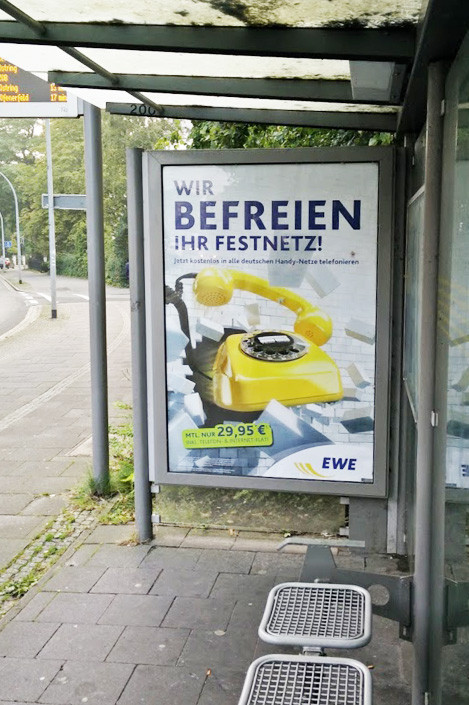 Frese & Wolff Werbeagentur, Citylight an einer Bushaltestelle für EWE, zu sehen ist ein gelbes Telefon, das eine Mauer durchbricht, dazu der Text "Wir befreien Ihr Festnetz, jetzt kostenlos in alle deutschen Handynetze telefonieren"