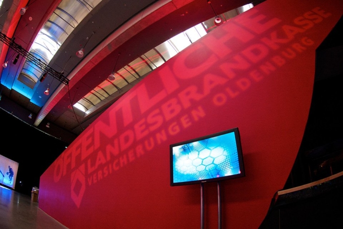 Frese & Wolff Werbeagentur, Foto vom Livemarketing für die Öffentliche Oldenburg, zu sehen ist eine rote Wand mit einem Fernseher davor